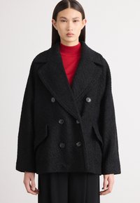 Melnā divrindas veste ar lieliem pogām, izgatavota no teksturētas auduma. Tam ir plats apkakle un sānu kabatas. Uzvilkta virs sarkana turtlenecka.