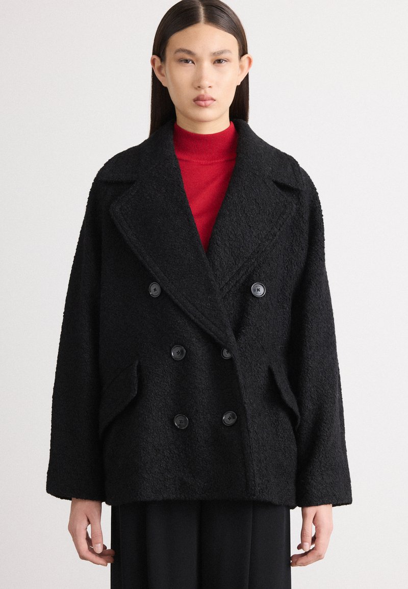 Melnā divrindas veste ar lieliem pogām, izgatavota no teksturētas auduma. Tam ir plats apkakle un sānu kabatas. Uzvilkta virs sarkana turtlenecka.