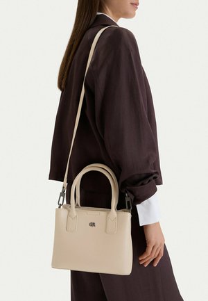 Mujer con un conjunto marrón oscuro llevando un bolso de cuero beige con asas dobles y una correa ajustable para el hombro.