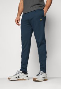 Navy blue sweatpants in a tapered design, made of soft fabric, featuring side pockets and a small yellow emblem. Worn with white sneakers.