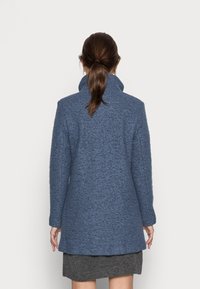 Modrý kabát z vlněné směsi s vysokým límcem, dlouhými rukávy a přímým střihem, s texturovaným povrchem a délkou sahající ke kolenům.