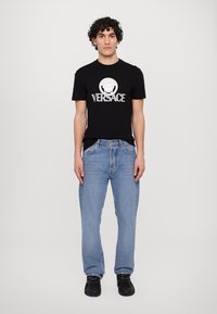 Sort bomulds T-shirt med hvid "VERSACE" logo og grafik, parret med lyseblå denim jeans. Modellen har sorte sko på.