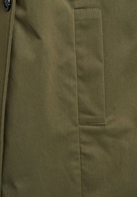 Tessuto verde oliva con una texture liscia, caratterizzato da una tasca laterale con dettagli di cucitura visibili. Il materiale appare durevole e strutturato.