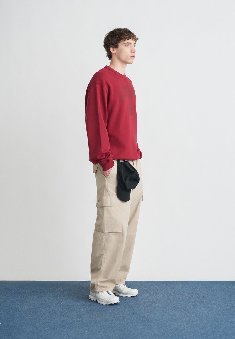 Sweat-shirt rouge à col rond, pantalon cargo beige, baskets blanches et casquette noire. Coupe décontractée avec coutures visibles et poches. Design simple.