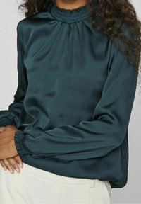 Bluzka z satyny w kolorze morskim, z marszczonym, wysokim kołnierzem i marszczonymi rękawami. Miękka, gładka w dotyku i o luźnym kroju. Noszona z jasnymi spodniami lub spódnicą.