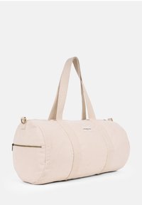 Round beige canvas duffel bag with two fabric straps, a front zip pocket, and a logo patch. Smooth texture with reinforced stitching.