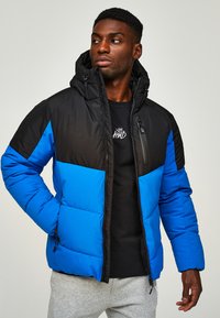 Blau- und schwarze Steppjacke mit Kapuze, ausgestattet mit einer gesteppten Textur, einem Frontreißverschluss und einer äußeren Reißverschlusstasche. Getragen über einem schwarzen Shirt.