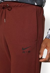 Bordeaux tepláky vyrobené z měkké látky, s černým logem Nike a textem "AIR", elastickým pasem a bočními kapsami.