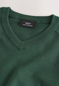Pull en maille vert foncé à encolure en V avec col côtelé et une étiquette noire portant l'inscription "NEXT Made in Bangladesh" à l'intérieur de l'encolure.