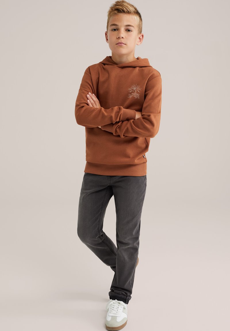Bruine hoodie met een grafische print, grijze spijkerbroek en witte sneakers. De hoodie heeft een kangoeroezak en ribgebreide manchetten. Gladde textuur.