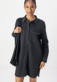 Black denim shirt dress with long sleeves, button front, and two chest pockets. Smooth texture and a relaxed fit, reaching mid-thigh.