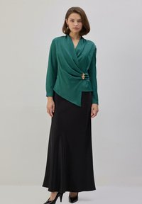 Mujer con blusa envolvente de manga larga en color verde esmeralda con broche dorado y falda larga negra, de pie contra un fondo liso y claro.