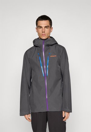 Man wearing a dark gray Patagonia hooded jacket with purple zipper and blue chest pocket zippers, standing against a plain background.