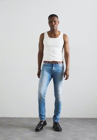 Camisa blanca sin mangas, jeans ajustados de color azul claro, cinturón de cuero marrón y zapatos negros, usados por una persona que está de pie contra un fondo liso.