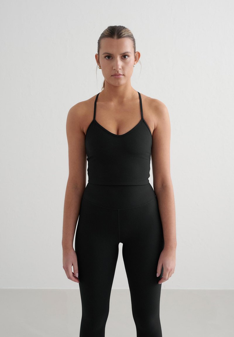 Zwarte geribde sportbeha met dunne bandjes, gecombineerd met hoge zwarte leggings. De gladde textuur en de aansluitende pasvorm creëren een samenhangende look.