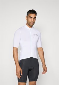 Maillot de cyclisme à manches courtes en blanc, avec une fermeture éclair à l'avant et un logo. Associé à un short de cyclisme noir rembourré. Design lisse et ajusté.