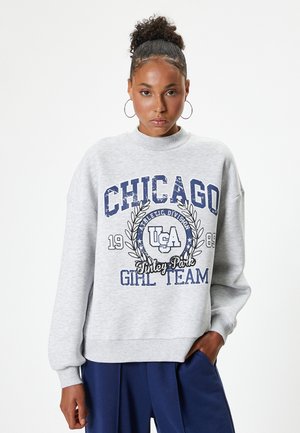 Sudadera gris con un cuello alto y un corte oversize. El texto gráfico azul dice "CHICAGO" y "GIRL TEAM" con toques atléticos y fechas.