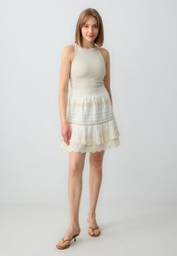 Débardeur en beige clair, associé à une jupe blanche à volants ornée de dentelle et de rayures horizontales. Des sandales à talons nude complètent le look.