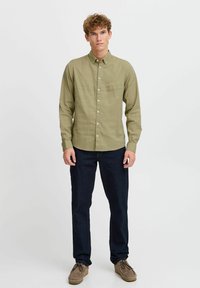 Cămașă lungă, verde măsliniu, cu guler cu butoane, textură verticală și logo subtil; purtată cu blugi negri și pantofi din suede maro.