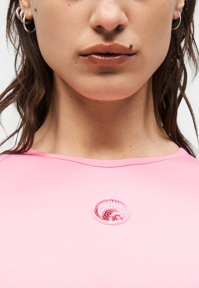 Κοντινό πλάνο στο κατώτερο μέρος του προσώπου και το λαιμό μιας γυναίκας που φοράει κρίκους στα αυτιά και ένα ροζ τοπ με κυκλικό σχέδιο από χάντρες κοντά στον γιακά.