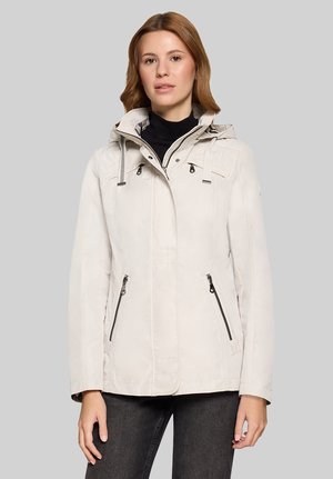 Frau trägt eine hellbeige Kapuzenjacke mit Reißverschlusstaschen und schwarze Hosen, steht vor einem schlichten grauen Hintergrund.