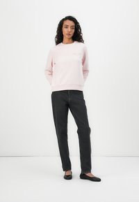 Lichtroze sweatshirt met een ronde hals en subtiele logo op de borst, gecombineerd met donkergrijze rechte broek en zwarte ballerina's.