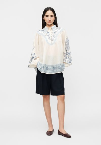 Γυναίκα που φοράει μια χαλαρή λευκή μπλούζα με μπλε λουλουδένια σχέδια, μαύρα σορτς μέχρι το γόνατο και σκούρα καφέ επίπεδα παπούτσια, στέκεται μπροστά σε ένα λευκό φόντο.