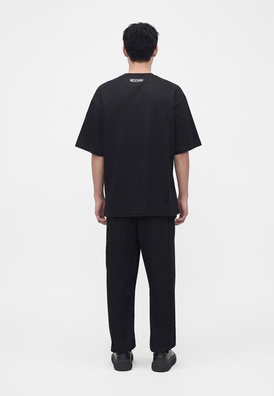 Homme portant un T-shirt noir Moschino oversize et un pantalon noir, debout dos à la caméra sur un fond blanc uni.