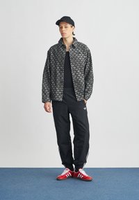 Fekete mintás dzseki galléros nyakkivágással fekete ing felett, fekete nadrággal, amelyen fehér csíkok vannak, valamint piros cipőkkel.