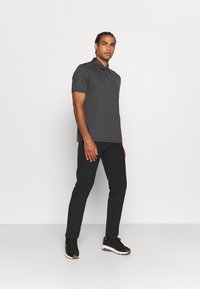 Homem de pé contra um fundo branco liso, vestindo uma camisa polo cinza escuro, calças slim pretas e ténis pretos com solas brancas.