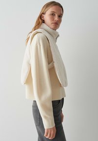 Gebreide crèmekleurige trui met een overlay van een nep-bonten vest. Het vest heeft een hoge kraag, lichte roze accenten en wordt open gedragen over de trui.
