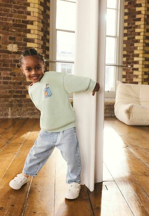 Ein kleines Kind lächelt und lehnt sich um eine weiße Säule in einem lichtdurchfluteten Raum mit Holzböden und freiliegenden Ziegelwänden.