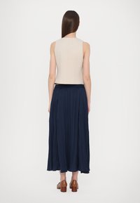 Beige top bez rukávů, kombinovaný s dlouhou tmavě modrou plisovanou sukní a hnědými botami na podpatku. Hladký textil s přiléhavým střihem.