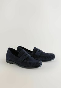 Paire de mocassins en daim bleu foncé avec un détail de bride et des semelles basses en caoutchouc noir sur une surface claire.