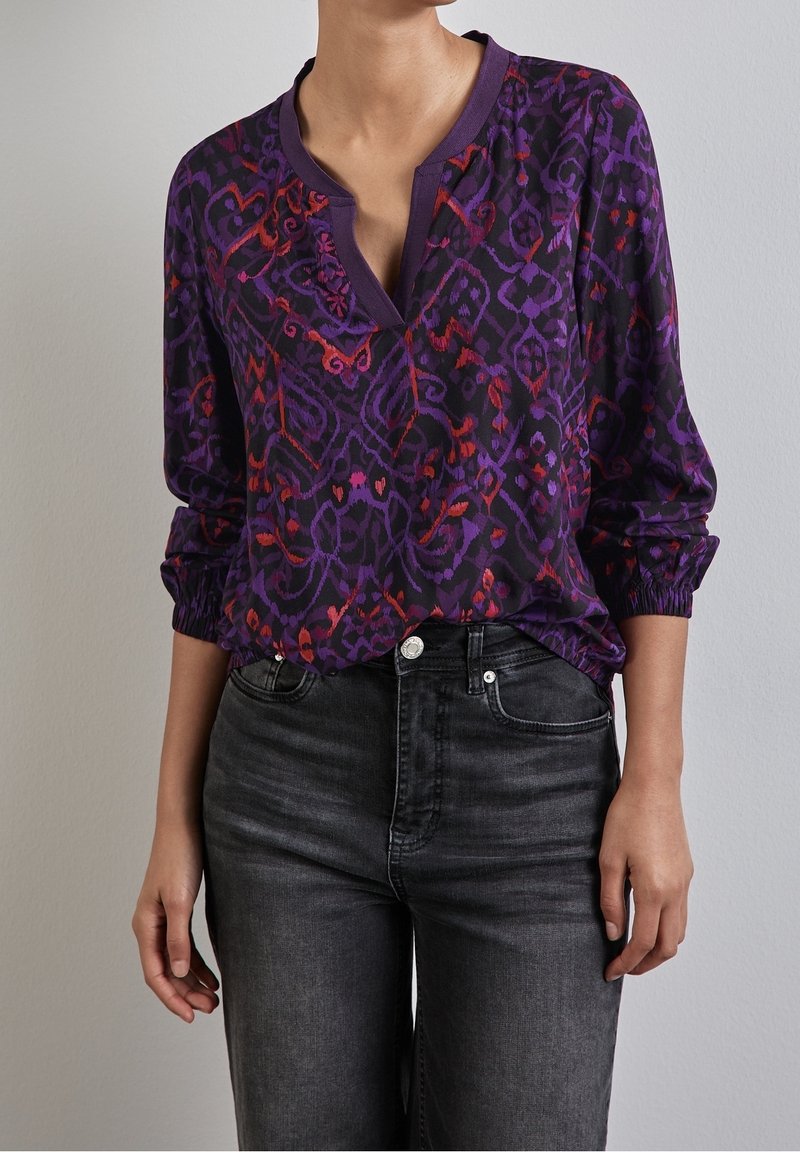 Blusa morada con estampado de diseños rojos y violetas, escote en V y detalle de botones; tela semitransparente con puños elásticos. Combinada con jeans negros.