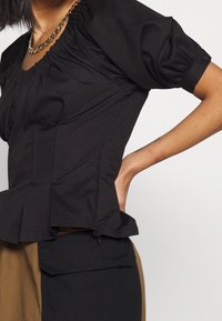 Schwarzes Oberteil mit Puffärmeln, gerafftem Ausschnitt, tailliertem Oberteil und Peplum-Saum. Aus glattem Stoff mit dezentem Glanz. Akzentuiert mit einer goldenen Kettenkette.