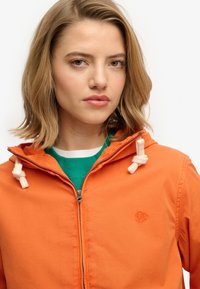 Giacca arancione con cerniera e cappuccio, caratterizzata da dettagli con coulisse crema e logo ricamato. Indossata sopra una maglietta verde a girocollo.