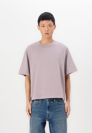 T-shirt lavande à manches courtes avec un col rond et une coupe décontractée, fabriquée en tissu coton doux, portée avec un jean droit bleu.