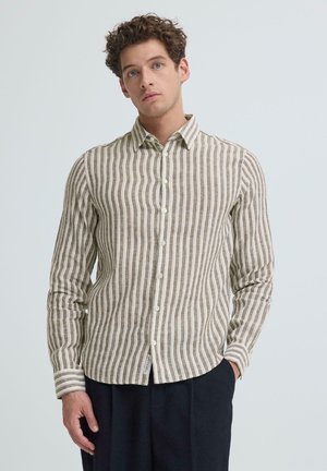 Young man wearing a beige and white vertical striped button-up shirt and dark pants, standing with one hand in pocket against plain background.