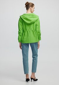 Groene lichtgewicht jas met capuchon, getailleerde taille, elastische manchetten, gecombineerd met lichtblauwe jeans en zwarte sleehakken.