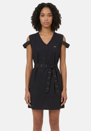 Donna che indossa un vestito nero senza maniche con scollo a V, spalline e cintura annodata in vita, in piedi davanti a uno sfondo chiaro e uniforme.