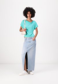 Top turchese con colletto ripiegato, abbinato a una gonna maxi in denim azzurra chiara con uno spacco laterale. Sneakers bianche completano l'outfit.