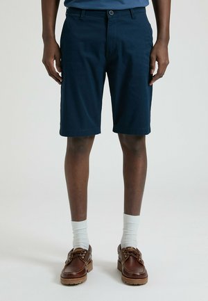 Pantaloncini in cotone blu marino con un taglio dritto, caratterizzati da chiusura con bottone e tasche laterali. Abbinati a scarpe di pelle marrone e calzini bianchi.