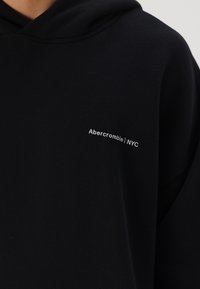 Hanorac negru cu o textură moale. Are un mic logo alb "Abercrombie | NYC" pe partea stângă a pieptului. Design casual.