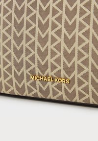 Brūna kanvas soma ar krēmīgu ģeometrisku rakstu. Zelta "MICHAEL KORS" logo tuvu apakšai. Gluda tekstūra ar strukturētu formu.