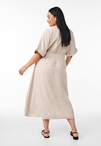 Beige linnklänning med korta uppvikt ärmar, åtsmitande midja och en flödande design som når nedanför anklarna. Sandaler kompletterar outfiten.