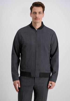 Mann trägt eine dunkelgraue Reißverschlussjacke mit schwarzem Kragen und Saum sowie passende graue Hose, steht vor einem schlichten hellen Hintergrund.