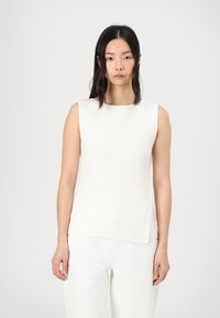 Top à côtes sans manches en blanc cassé avec un col rond et des boutons sur le côté. Le tissu a une apparence douce et texturée.