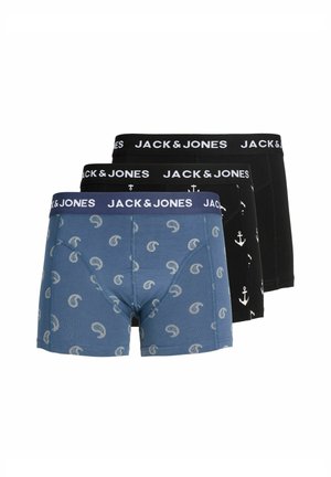 Mladý muž s krátkymi blond vlasmi, oblečený v modrých vzorovaných boxerkách s pásom s logom Jack & Jones, stojaci pred jednoduchým svetlým pozadím.