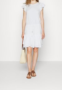 White knee-length dress with ruffled cap sleeves, a drawstring waist, and scalloped hem. Paired with brown sandals and a woven bag.